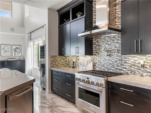 kitchen with backsplash, wall chimney range hood, light countertops, appliances with stainless steel finishes, and marble finish floor