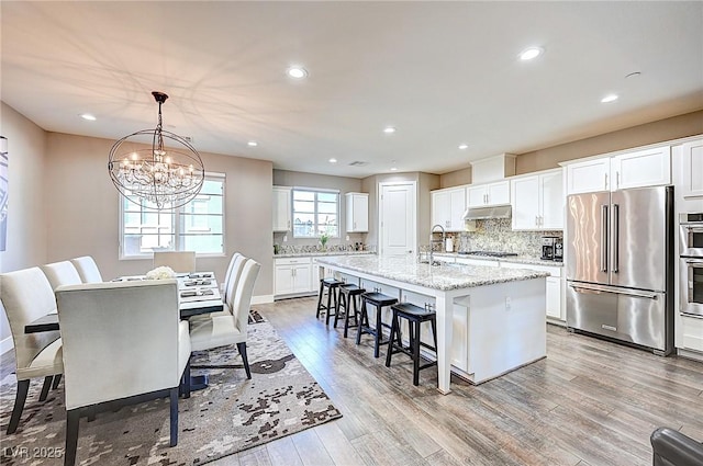 kitchen with light wood-style flooring, appliances with stainless steel finishes, a kitchen breakfast bar, under cabinet range hood, and backsplash