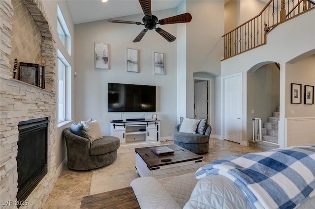 living area with ceiling fan, stairway, a stone fireplace, a towering ceiling, and arched walkways