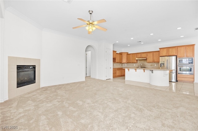 unfurnished living room featuring crown molding, a tiled fireplace, light colored carpet, arched walkways, and a ceiling fan