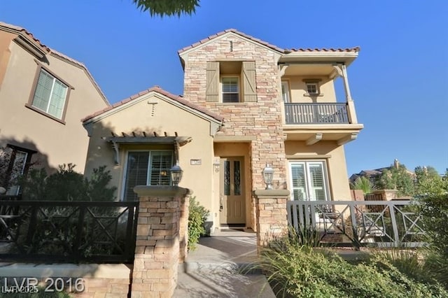 mediterranean / spanish-style house with fence, a tiled roof, stucco siding, a balcony, and stone siding