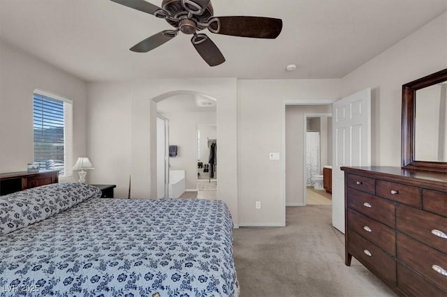 bedroom featuring a ceiling fan, ensuite bath, arched walkways, baseboards, and light colored carpet