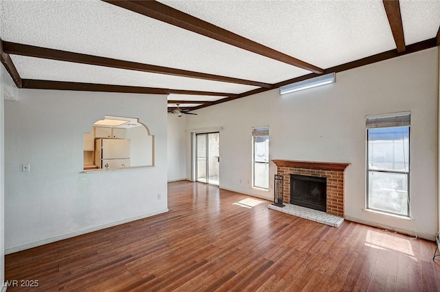 unfurnished living room featuring beamed ceiling, a textured ceiling, wood finished floors, a brick fireplace, and ceiling fan