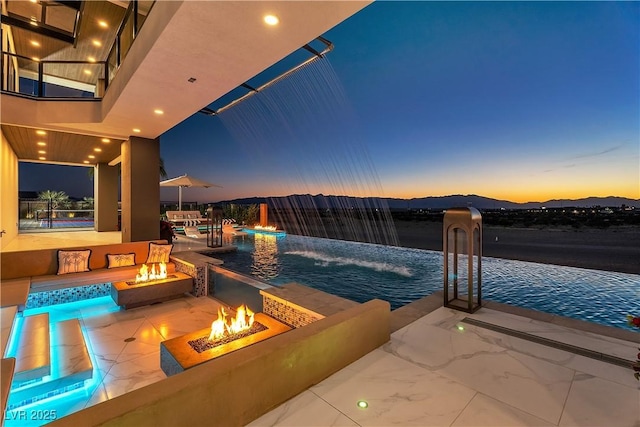 pool at dusk with a patio area, an infinity pool, a mountain view, and an outdoor fire pit