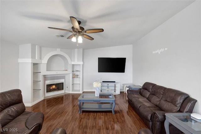 living area featuring visible vents, a warm lit fireplace, wood finished floors, baseboards, and ceiling fan