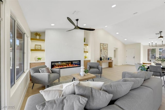 living area with a ceiling fan, visible vents, vaulted ceiling, a glass covered fireplace, and light wood-type flooring