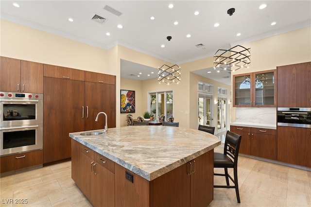 kitchen with visible vents, glass insert cabinets, double oven, an island with sink, and a sink