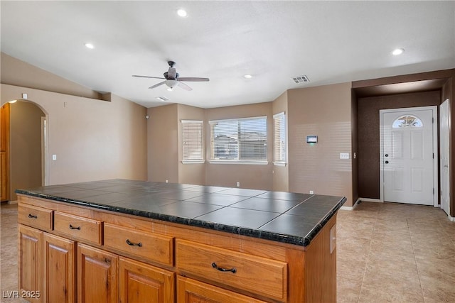 kitchen featuring tile countertops, arched walkways, brown cabinets, and ceiling fan