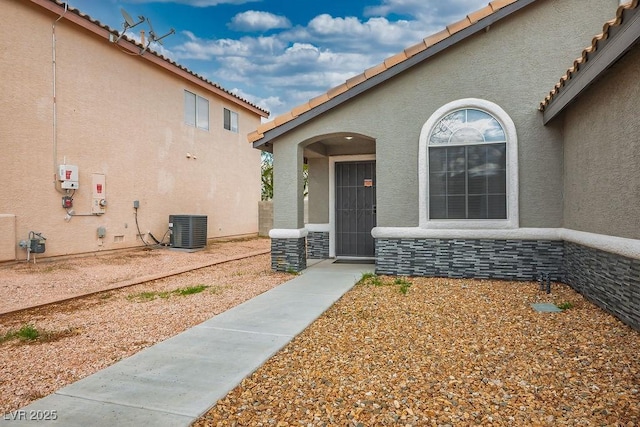 doorway to property with stone siding, central AC unit, a tile roof, and stucco siding