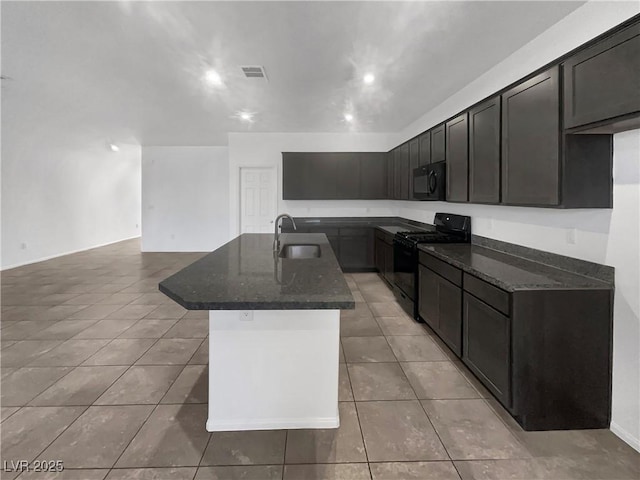 kitchen with light tile patterned floors, dark stone counters, an island with sink, a sink, and black appliances