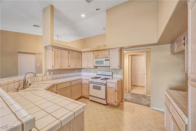 kitchen featuring light brown cabinetry, a sink, white appliances, a peninsula, and tile counters