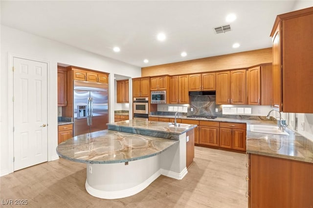 kitchen featuring a sink, light stone countertops, built in appliances, brown cabinetry, and a kitchen island with sink