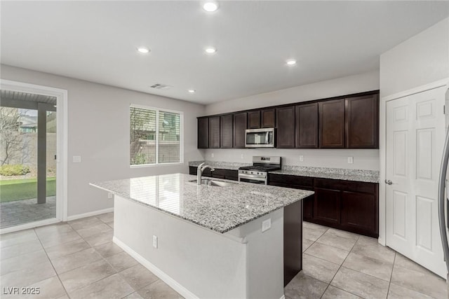 kitchen with dark brown cabinetry, an island with sink, light stone counters, appliances with stainless steel finishes, and a sink