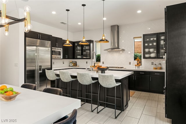 kitchen featuring wall chimney range hood, built in appliances, dark cabinets, a breakfast bar area, and a sink