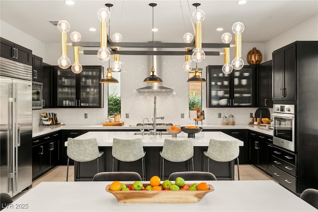 kitchen with built in appliances, dark cabinets, a breakfast bar area, light countertops, and a kitchen island with sink