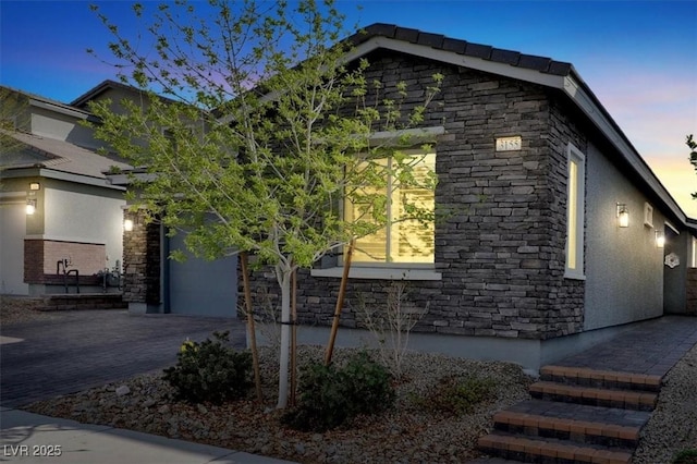 property exterior at dusk featuring stucco siding, stone siding, a garage, and driveway