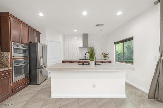 kitchen featuring visible vents, an island with sink, light countertops, stainless steel appliances, and wall chimney exhaust hood