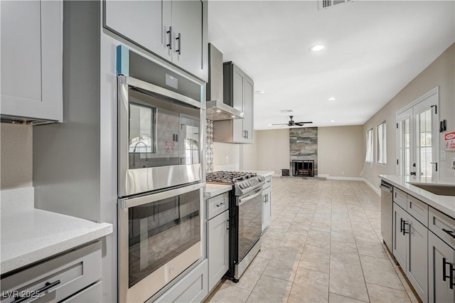 kitchen featuring gray cabinets, stainless steel appliances, wall chimney range hood, open floor plan, and a large fireplace