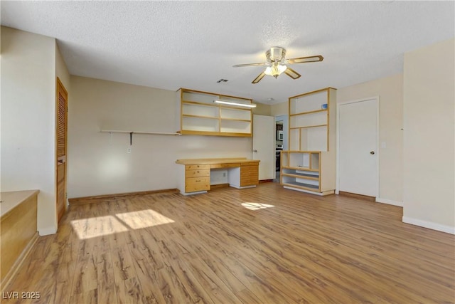 unfurnished living room with ceiling fan, light wood-style floors, built in desk, and a textured ceiling