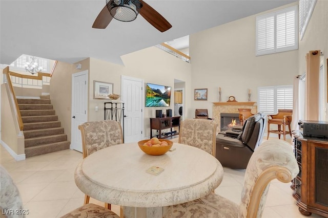 dining area with stairway, light tile patterned floors, a warm lit fireplace, a high ceiling, and a ceiling fan