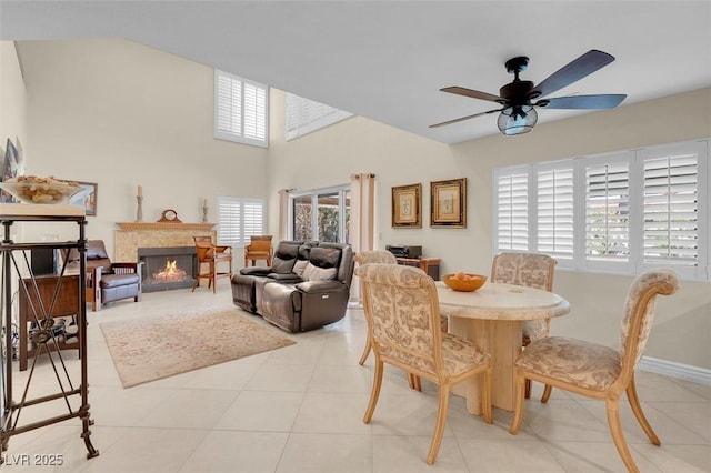 dining space with a ceiling fan, baseboards, a warm lit fireplace, and light tile patterned floors