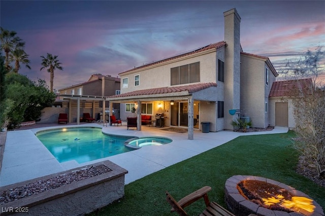 back of house at dusk with a lawn, a pool with connected hot tub, a fire pit, and a patio