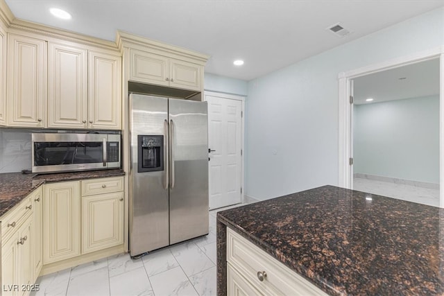 kitchen with dark stone countertops, visible vents, stainless steel appliances, cream cabinetry, and marble finish floor