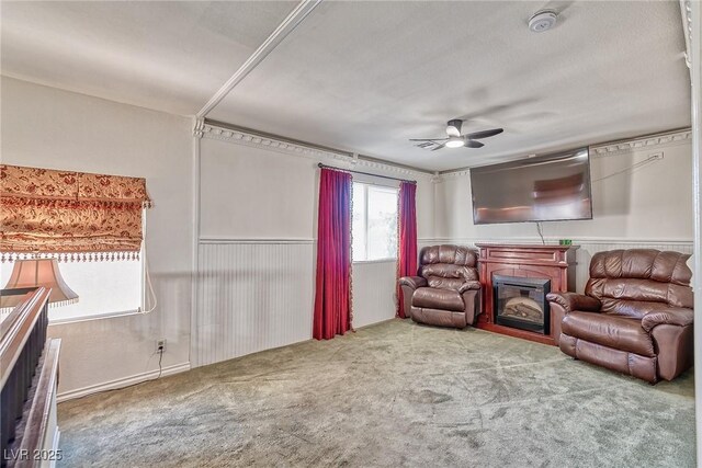 carpeted living room featuring a glass covered fireplace, a ceiling fan, and wainscoting