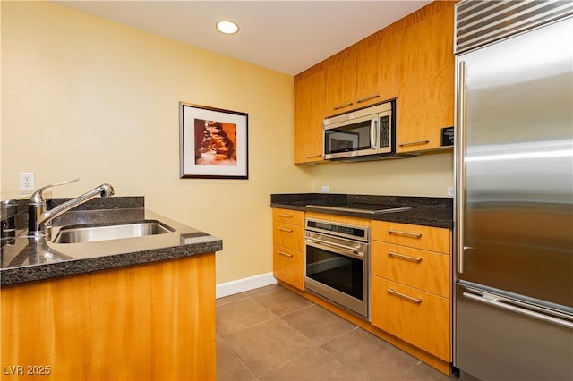 kitchen with dark stone counters, dark tile patterned flooring, a sink, appliances with stainless steel finishes, and brown cabinets