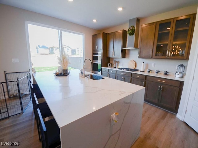 kitchen with an island with sink, light stone counters, wood finished floors, appliances with stainless steel finishes, and wall chimney range hood