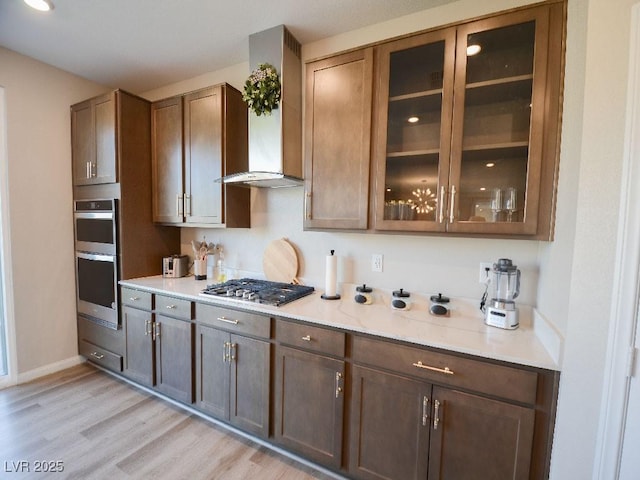 kitchen with glass insert cabinets, baseboards, wall chimney range hood, light wood-type flooring, and appliances with stainless steel finishes