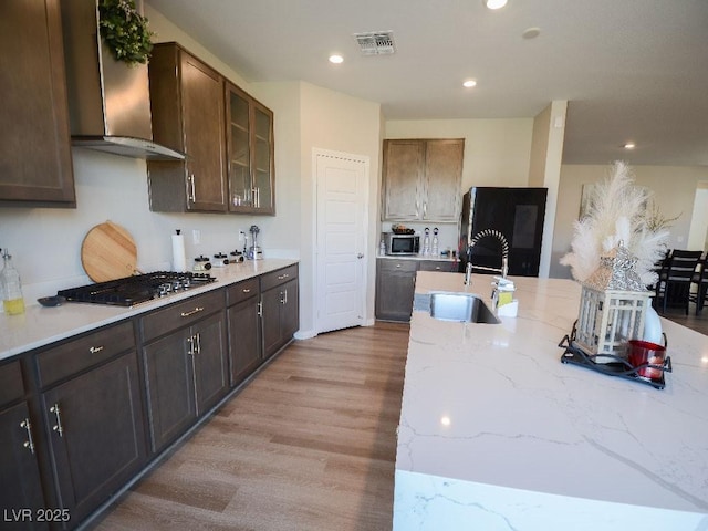 kitchen featuring a sink, glass insert cabinets, light wood-style floors, appliances with stainless steel finishes, and wall chimney exhaust hood