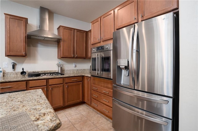 kitchen featuring light stone counters, light tile patterned floors, stainless steel appliances, wall chimney exhaust hood, and brown cabinets