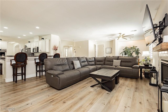 living area featuring visible vents, light wood-style floors, arched walkways, a glass covered fireplace, and a ceiling fan