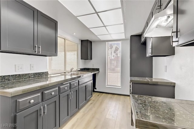 kitchen with gray cabinetry, tile counters, under cabinet range hood, light wood-style flooring, and a sink