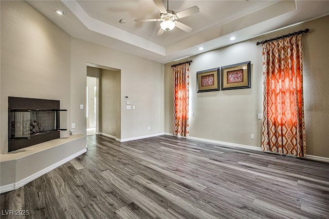 unfurnished living room featuring wood finished floors, baseboards, a tray ceiling, recessed lighting, and a fireplace with raised hearth