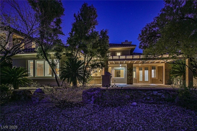 back of house at night with french doors, a chimney, and stucco siding