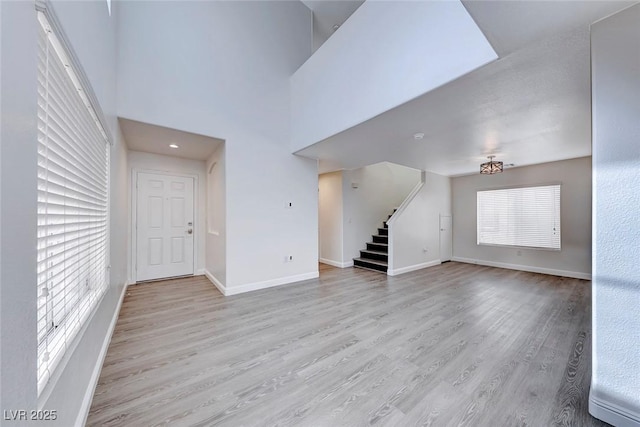 unfurnished living room featuring stairs, light wood-style flooring, baseboards, and a towering ceiling