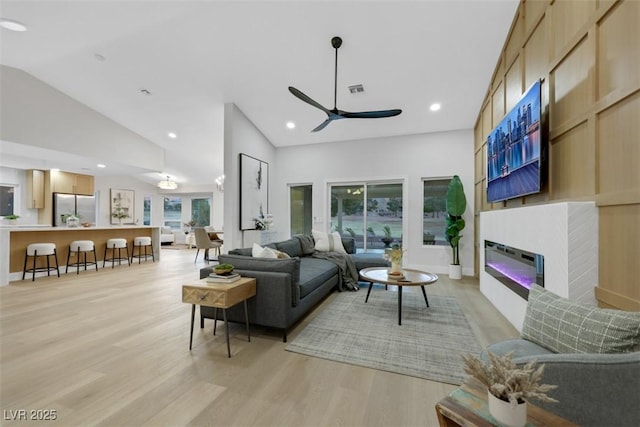 living room with visible vents, high vaulted ceiling, a glass covered fireplace, light wood-style floors, and ceiling fan