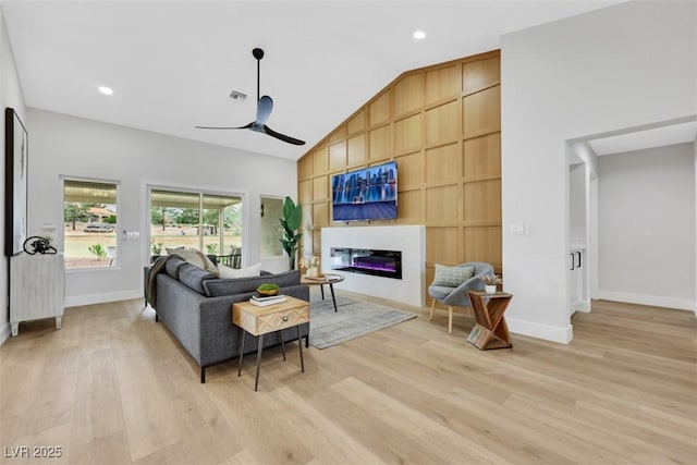 living room featuring light wood-type flooring, high vaulted ceiling, a glass covered fireplace, baseboards, and ceiling fan