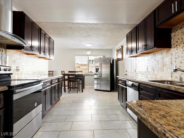 kitchen featuring light tile patterned floors, a sink, dark brown cabinetry, appliances with stainless steel finishes, and wall chimney exhaust hood