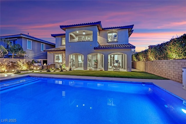 back of property at dusk with a balcony, a fenced in pool, stucco siding, a patio area, and a lawn