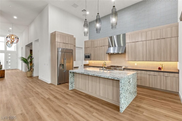 kitchen featuring light brown cabinetry, built in appliances, modern cabinets, and wall chimney range hood