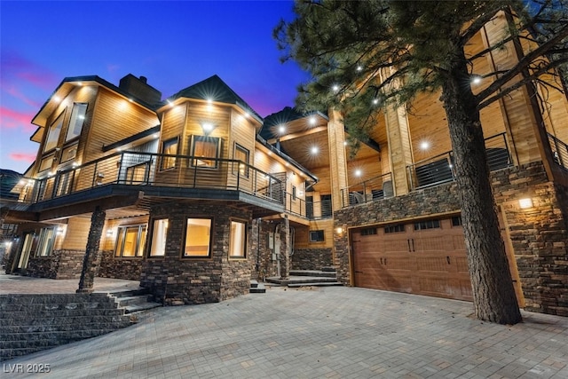 modern home with decorative driveway, stone siding, a balcony, and an attached garage