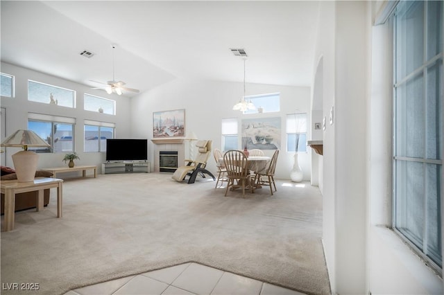 carpeted living room with visible vents, high vaulted ceiling, a tiled fireplace, and ceiling fan with notable chandelier