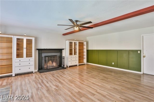 unfurnished living room with a fireplace with raised hearth, a wainscoted wall, beam ceiling, wood finished floors, and a ceiling fan