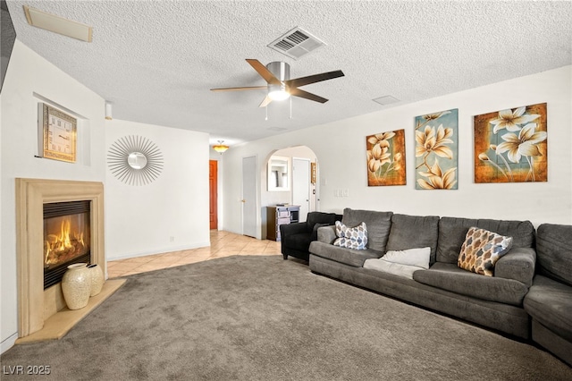 carpeted living room with tile patterned floors, visible vents, a glass covered fireplace, arched walkways, and ceiling fan