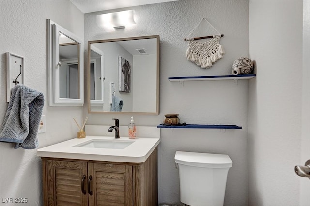 half bath featuring visible vents, toilet, vanity, and a textured wall