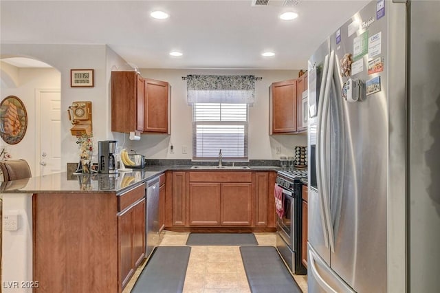 kitchen featuring brown cabinetry, a peninsula, arched walkways, a sink, and appliances with stainless steel finishes