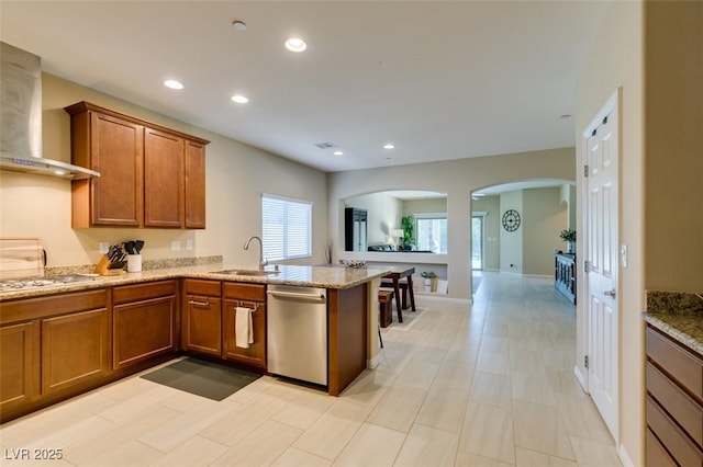 kitchen with wall chimney range hood, recessed lighting, brown cabinets, appliances with stainless steel finishes, and a sink